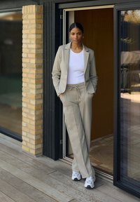 Completo beige chiaro con giacca aderente e pantaloni con coulisse. T-shirt bianca sotto. Sneakers casual completano l'outfit. Sfondo in legno e mattoni.