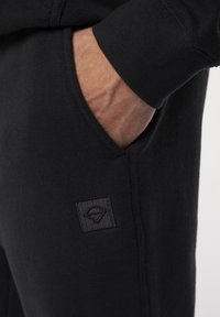 Svarta sweatpants med en texturerad tyg, elastiska mudd och en liten svart fyrkantig logotyp på fickan.