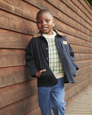 Νεαρός αγόρι με μπλε μπουφάν, πουλόβερ καρό και τζιν, ακουμπάει σε ξύλινο τοίχο, χαμογελώντας με τα χέρια στις τσέπες σε εξωτερικό χώρο.