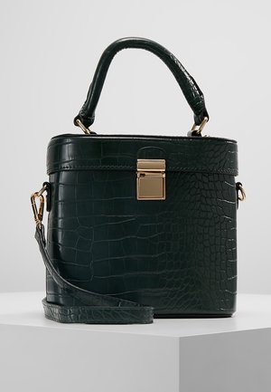 Ciemnozielona torebka z wzorem krokodyla, z górnym uchwytem, złotym zapięciem i odpinanym paskiem na ramię, na białym piedestale.
