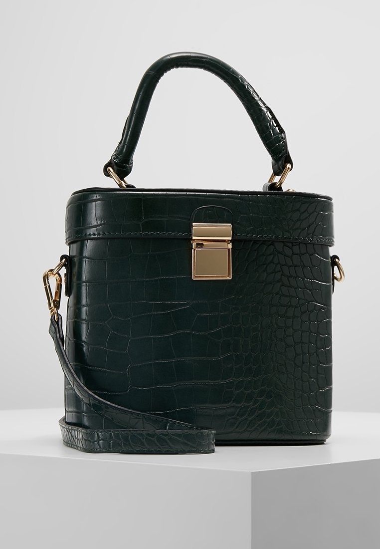Sac à main vert foncé à motif crocodile avec poignée supérieure, fermoir doré et bandoulière amovible sur un piédestal blanc.