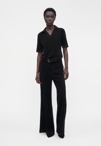 Femme debout portant une chemise noire à manches courtes avec col, un pantalon large noir ceinturé et des chaussures noires pointues sur fond blanc.