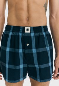 Miehekäs torso, jossa on tummansinisiä ja vaaleansinisiä ruudullisia boxereita, joissa on joustava vyötärönauha ja nappisuljenta, sekä osittain näkyvä tatuointi oikealla puolella.