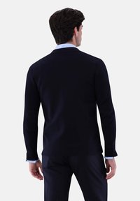 Marineblauer Pullover mit langen Ärmeln, gerippten Bündchen und einer glatten Textur; auffällige Ellbogenpatches, getragen über einem hellblauen Hemd mit Kragen.