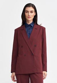 Kobieta w bordowej marynarce dwurzędowej i spodniach oraz w granatowej koszuli z guzikami, stojąca na tle jasnej, jednolitej ściany.