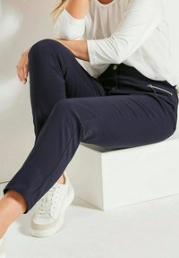 Marineblauwe stretchbroek met een slanke pasvorm, voorzien van een voorzak met rits en een soepele stof, gecombineerd met witte sneakers en een lichte top.