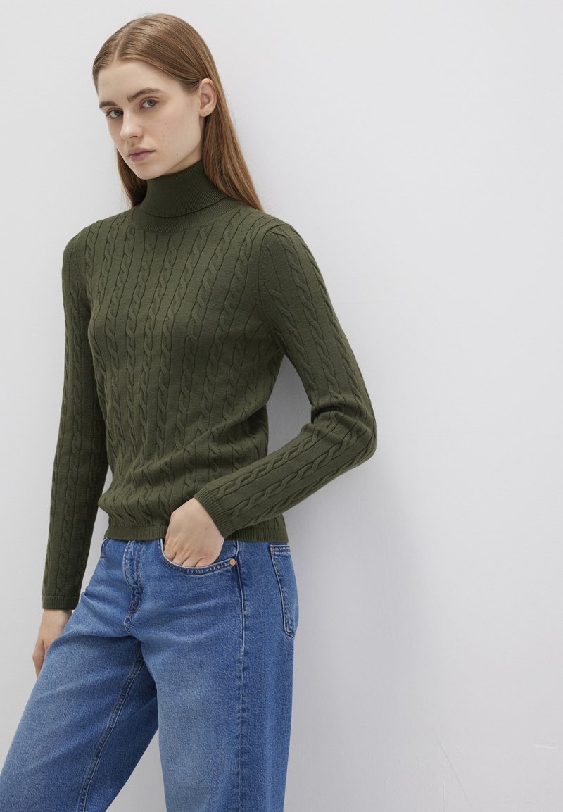 Olivgrüner Strickrollkragenpullover mit einem Zopfmuster und gerippten Bündchen. Kombiniert mit hellblauen, lässigen Jeans mit einer Fronttasche.
