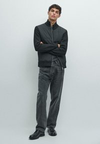 Giacca nera con zip e pannello superiore con texture, abbinata a jeans grigi a gamba larga e scarpe nere, con vestibilità comoda e collo alto.