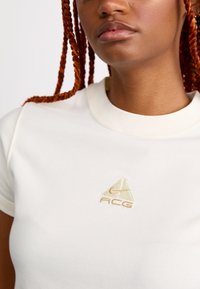 Nærbilde av en person med røde fletter som har på seg en hvit Nike ACG t-skjorte, med fokus på brystlogoen og nedre del av ansiktet.