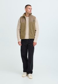 Fleece vest in tan with a textured lower panel, paired with a cream cable-knit sweater and black jeans. Footwear is light-colored sneakers.