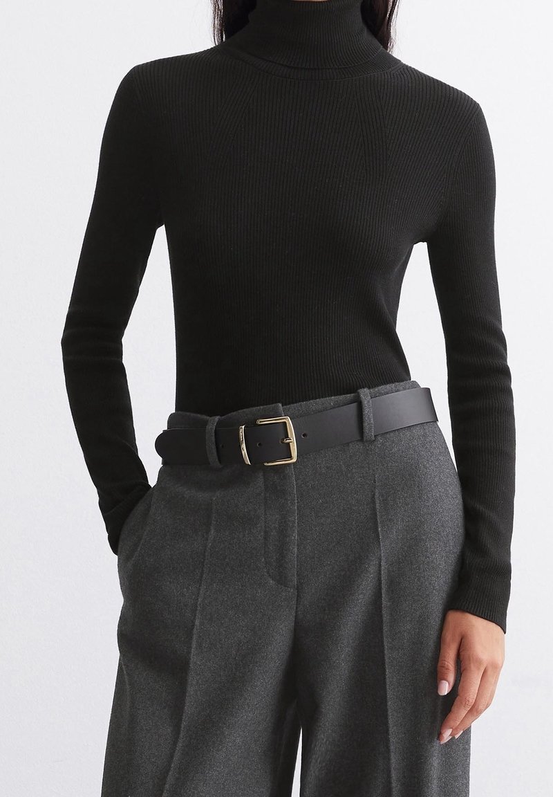 Pull à col roulé noir côtelé associé à un pantalon gris taille haute et une ceinture noire avec une boucle dorée. Texture de tissu lisse.