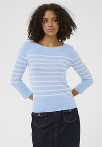 Femme aux cheveux bouclés portant un pull bleu clair avec des rayures blanches horizontales et une jupe en denim foncé, se tenant les mains derrière le dos.