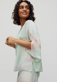 Helle, pastellgrüne Bluse mit Aquarellmuster, weiten Ärmeln und seitlichen Schlitzen. Hergestellt aus leichtem, durchscheinendem Stoff mit einer weichen Textur.