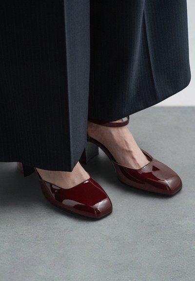 Escarpins en cuir verni bordeaux avec un design à bout carré, dotés d'une fine bride à la cheville et d'un épais talon bloc. Pantalon en tissu texturé visible.