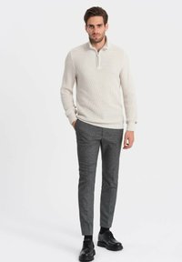 Pull blanc en maille texturée avec un col zippé moitié, associé à un pantalon gris ajusté à motifs et des chaussures noires à lacets.
