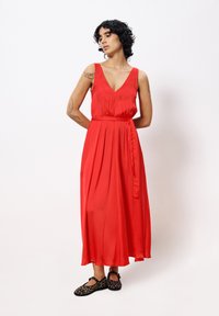 Red, sleeveless maxi dress with a deep V-neck, pleated bodice, and a waist tie. Melon shape, flowing fabric, paired with black leopard-print shoes.