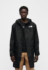 Μακρύ αδιάβροχο μαύρο παλτό με κουκούλα, με εμπρόσθιο κλείσιμο με κουμπιά, δύο εξωτερικές τσέπες και λογότυπα στο στήθος.