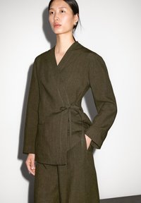 Olijfgroene wikkel-top van gestructureerde stof; heeft een zijstrik, lange mouwen en een V-halslijn, gecombineerd met bijpassende broek.