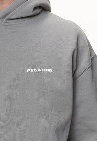 Felpa grigia realizzata in tessuto morbido, con tasca a marsupio, polsini a coste e logo bianco "PEGADOR" sul petto.