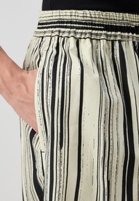 Løse bukser i kremfarge med vertikale svarte striper, med elastisk midje og sidelommer. Glatt tekstur med lett design.