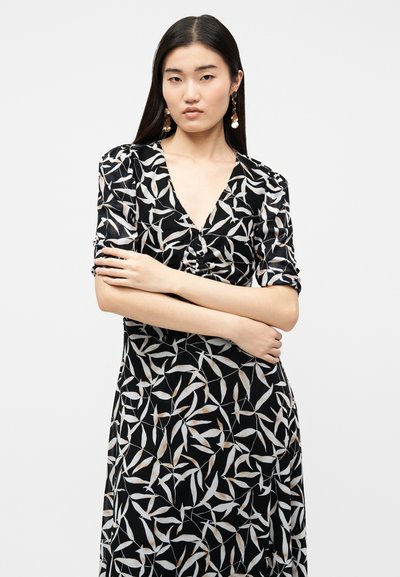 Γυναίκα με μακριά σκούρα μαλλιά, που φορά μαύρο φόρεμα με μπεζ σχέδιο φύλλων και χρυσά κρεμαστά σκουλαρίκια, με τα χέρια σταυρωμένα μπροστά σε λευκό φόντο.