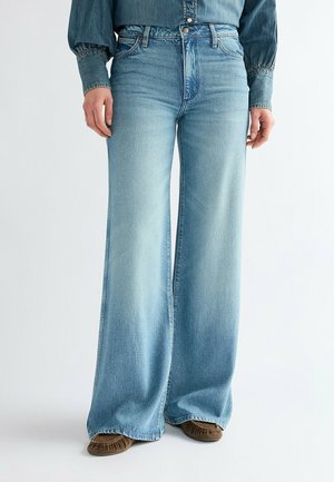 Persona che indossa jeans azzurri chiaro a vita alta e gamba larga, camicia di jeans infilata e mocassini marroni, in piedi davanti a uno sfondo semplice.