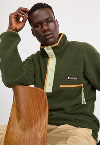 Grøn fleece-pullover med en lynlås-lomme, cremet knaplukning og orange detaljer. Stoffet er struktureret og blødt, hvilket giver varme.