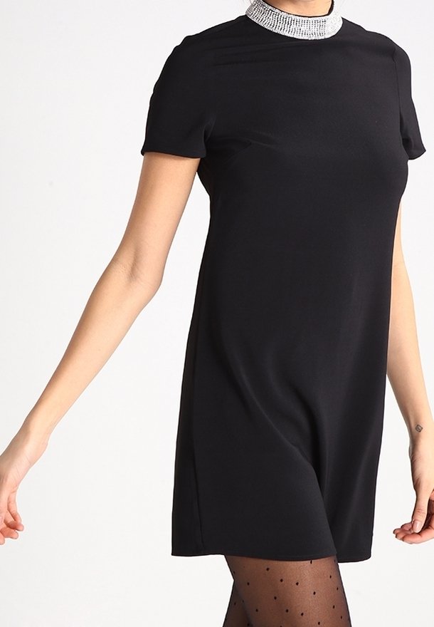 Femme portant une robe noire courte avec un col pailleté argenté et des collants noirs transparents à pois, debout devant un fond uni.