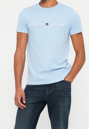 Homme portant un t-shirt bleu clair Tommy Hilfiger avec logo et un jean bleu foncé, une main dans la poche, sur fond blanc uni.