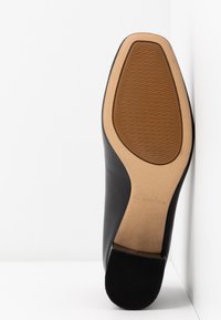 Semelle d'une chaussure habillée en cuir noir avec une semelle en caoutchouc marron texturée et beige, inclinée contre un coin blanc en arrière-plan.