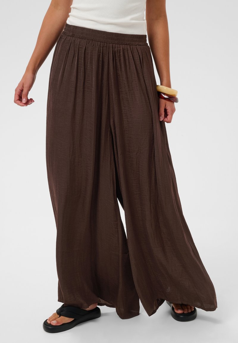Femme portant un pantalon large ample marron foncé, un top blanc sans manches, des sandales noires et deux bracelets épais au poignet droit.