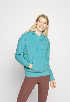 Hoodie turquesa com um bolso frontal, cordões e punhos canelados. Usado com leggings castanhas, apresentando um pequeno logótipo no peito.