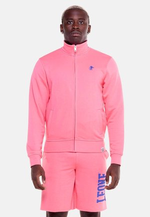 Man draagt een felroze jas met rits en bijpassende korte broek met blauwe "LEONE" tekst op de korte broek, tegen een effen achtergrond.