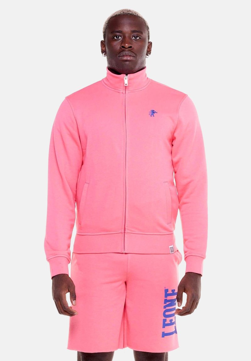 Homme portant une veste zippée rose vif et un short assorti avec le texte bleu « LEONE » sur le short, sur un fond uni.