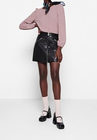 Langärmlige, mauvefarbene Bluse mit gerafften Schultern; schwarzer Leder-Minirock mit Reißverschluss vorne; blassrosa Socken; klobige schwarze Absatzschuhe.