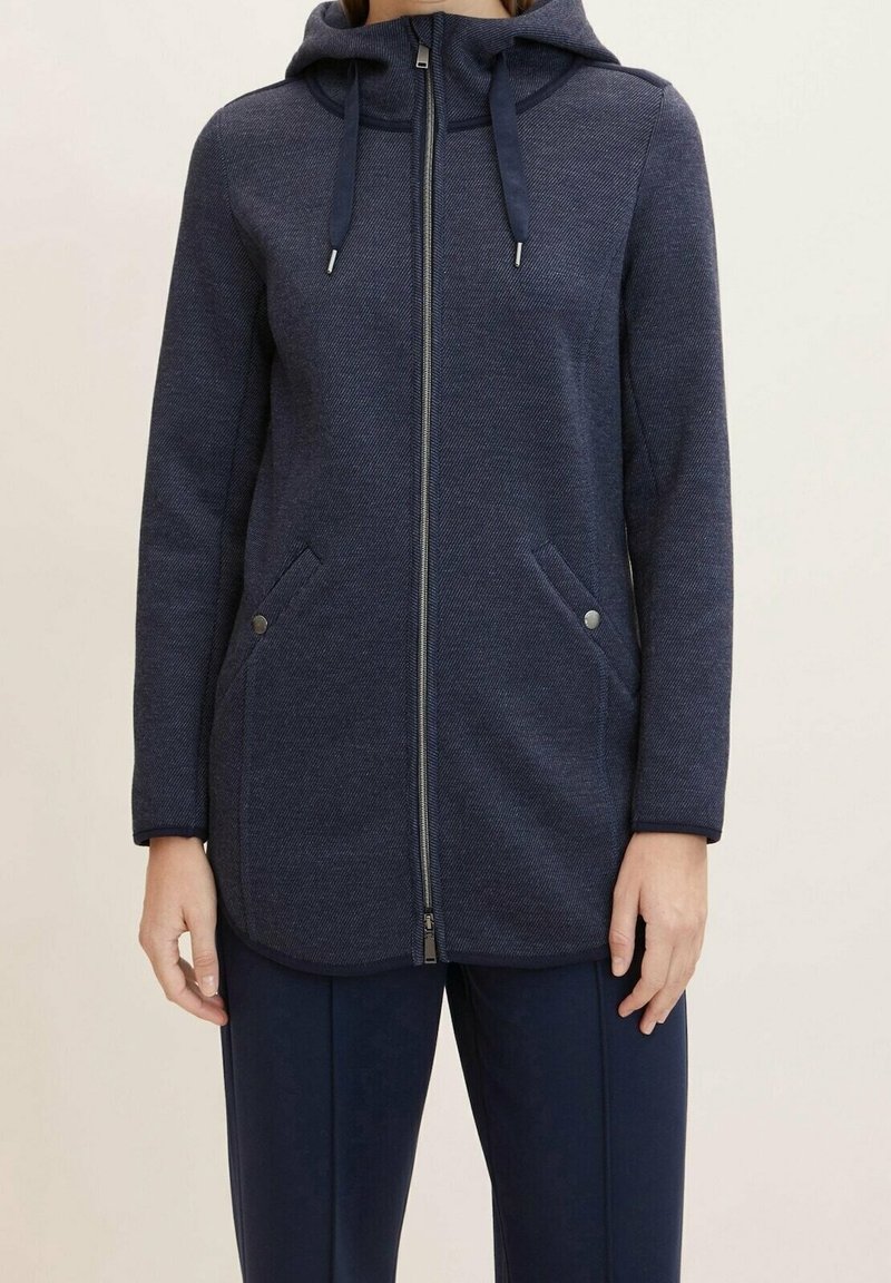 Personne portant une veste à capuche bleu marine zippée, à manches longues et avec des poches latérales, assortie à un pantalon bleu marine.