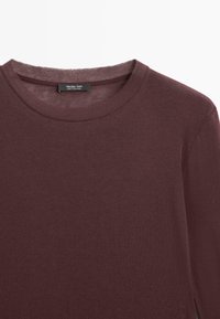 Burgunderfarbendes Baumwollhemd mit langen Ärmeln und Rundhalsausschnitt, ausgestattet mit einer weichen Textur und einem Etikett am inneren Ausschnitt, das den Markennamen zeigt.