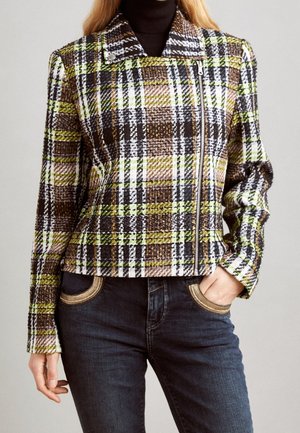 Žena nosící zelenou, hnědou, bílou a černou károvanou bundu s asymetrickým zipem přes černý rolák a tmavé džíny.