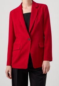 Červený blazer s jedním knoflíkem, klopovými límci, dvěma předními kapsami a hladkým materiálem; kombinovaný s černým topem a kalhotami.