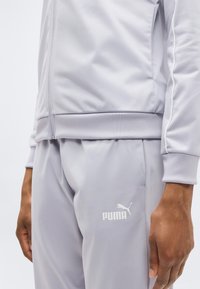 Ανοιχτό γκρι φόρμα από λείο ύφασμα, που περιλαμβάνει ζακέτα με φερμουάρ και παντελόνι με ελαστική μέση. Διακριτικό λευκό λογότυπο Puma στο αριστερό μπούτι.