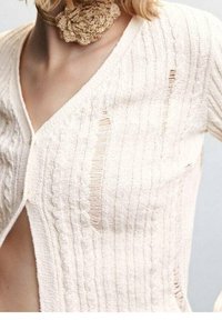 Creme-farbene Strickjacke mit vertikalen Rippen, die dezente Ausschnitte und ein V-Ausschnitt-Design aufweist. Weiche Textur, leichtes Material.