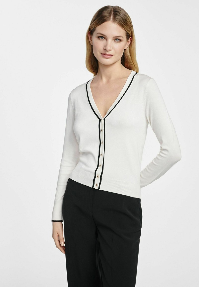 Biała kardigan z czarnym wykończeniem, dekoltem w serek i zapięciem na siedem guzików. Długie rękawy, dopasowany krój i ryflem o fakturze.