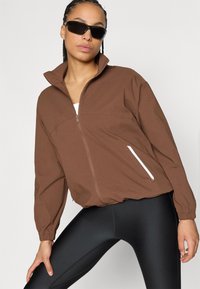 Καφέ ζακέτα με φερμουάρ, ψηλό γιακά και ελαστικά μανίκια, με πλαϊνές τσέπες και λευκές λεπτομέρειες. Φοριέται με μαύρο κολάν.