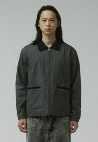 Chaqueta gris con cremallera y cuello negro, con dos bolsillos delanteros y un acabado ligeramente texturizado. Llevada sobre pantalones a juego de color gris claro.