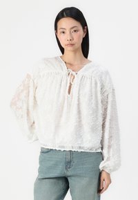 Blusa branca com textura floral, decote redondo e detalhe de laço. Apresenta mangas longas e um corte descontraído. Usada com jeans azuis.