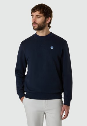 Homme barbu portant un sweat-shirt bleu marine et un pantalon gris clair, regardant vers la gauche avec les mains dans les poches, sur un fond uni.