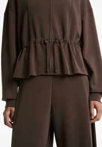 Brauner Zip-up Sweatshirt im Peplum-Stil mit Kordelzug-Taille, weichem Material und langen Ärmeln, kombiniert mit passenden Hosen.