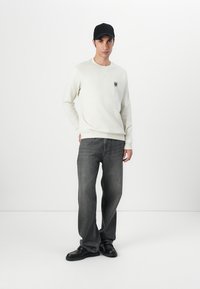 Hvit genser med logo, grå jeans, og svart caps. Genseren har rund hals og ribbede mansjetter; jeansene har en avslappet passform.