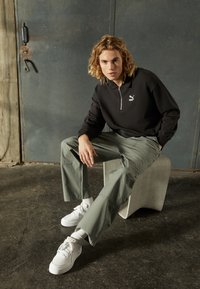 Czarna polo z zamkiem błyskawicznym i białym logo, zielone spodnie cargo i białe sneakersy. Model siedzi na teksturowanym jasnoszarym stołku na tle ciemnym.