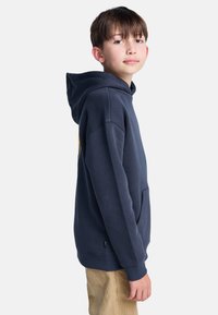 Granatowa bluza z kapturem z przednią kieszenią, miękką fakturą i ściągaczami na rękawach. Standardowy krój z obszernym kapturem. Nosi się ją na beżowe spodnie.